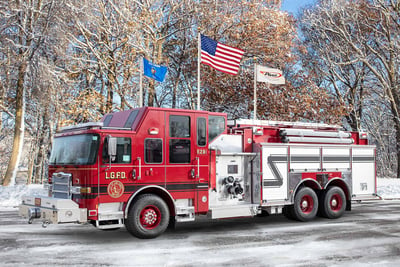 A red Pierce custom dry side tanker with an elevated cab and chrome finishes is parked in front of a snowy forest background with three flags above it. 