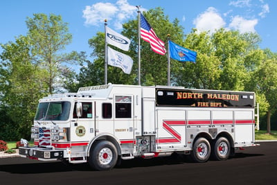 A white Pierce PUC tanker with a red stripe is parked in front of green trees with four flags flying overhead.  