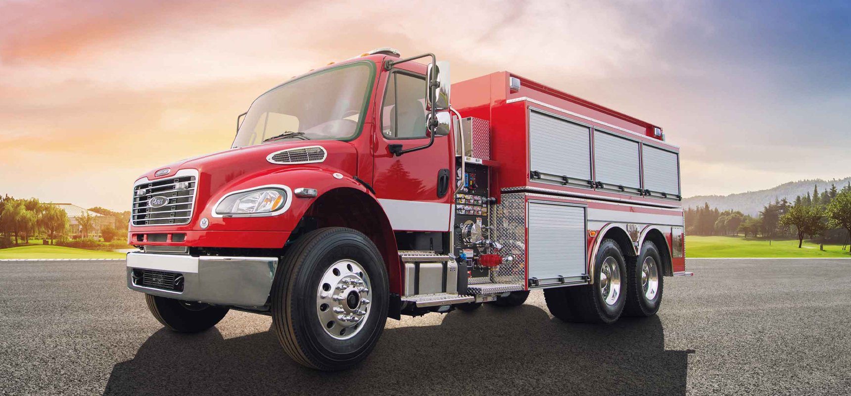 A red tanker fire truck with chrome finishes is parked in a lot with green foliage and low mountains in the background and a colorful sunset sky.  