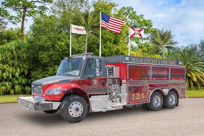 A red and gray Pierce BX tanker fire truck is parked with green foliage in the background below three waving flags.  