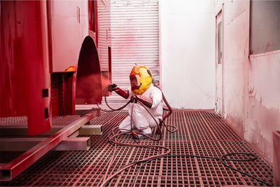 A person in a white paining suite and yellow protective cap in a speciality room painting a fire truck red.