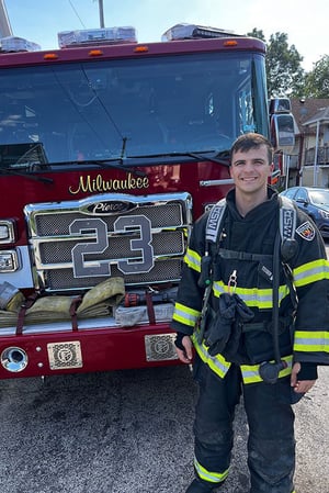 A male firefighter in uniform stands in front of a firetruck with a number 23 on the grill.