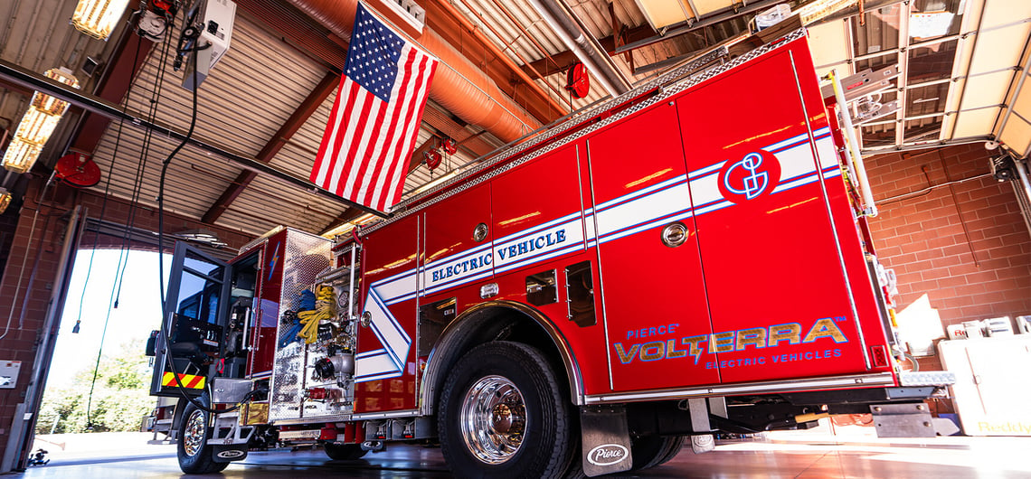 The red Pierce Volterra electric fire truck pumper is parked in a station bay with an American flag overhead after the push in ceremony. 