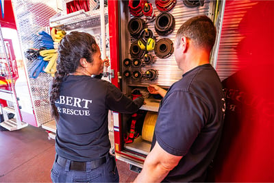 Two Gilbert Fire & Rescue firefighters are talking and working on compartmentation on the side of a red electric pumper fire truck.