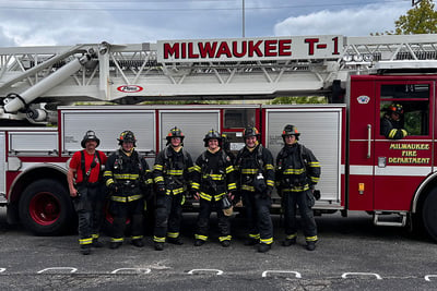 A group of firefighter's in uniform stand in front of a red fire truck with 'Milwaukee' written on the ladder.