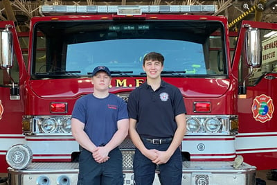 Two firefighter interns stand in front of a red fire truck.