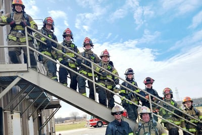 A group of fire fighter interns standing on a metal staircase with blue skies and white clouds in the background.