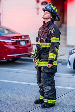 A firefighter in uniform looks up while standing on a street with cars in the background.