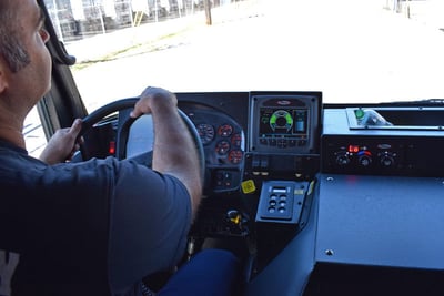 An over-the-shoulder image of a fire truck operator holding the steering wheel with the dashboard display and front window in view. 