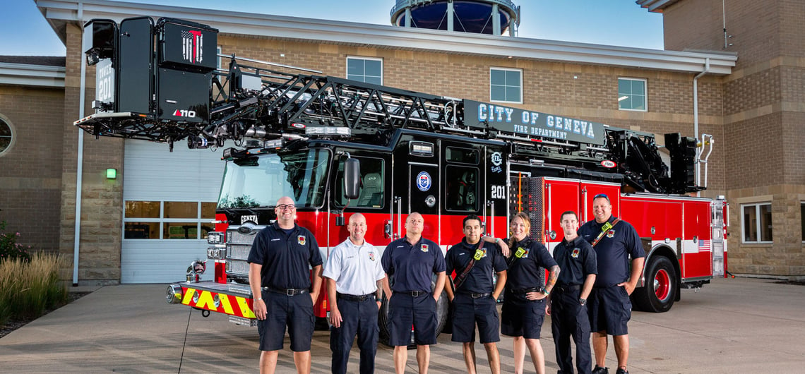 Seven firefighters stand in front of an aerial apparatus parked outside the City of Geneva fire station. 