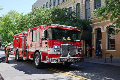 A red electric fire truck is driving down a city street with brick buildings and trees in the background.