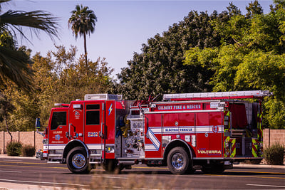 A red electric fire apparatus is driving down an asphalt road with a palm tree and other greenery in the background. 