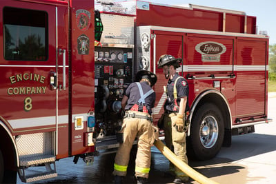 Two firefighters are connecting a hose to the side panel of a stationary red electric fire truck. 