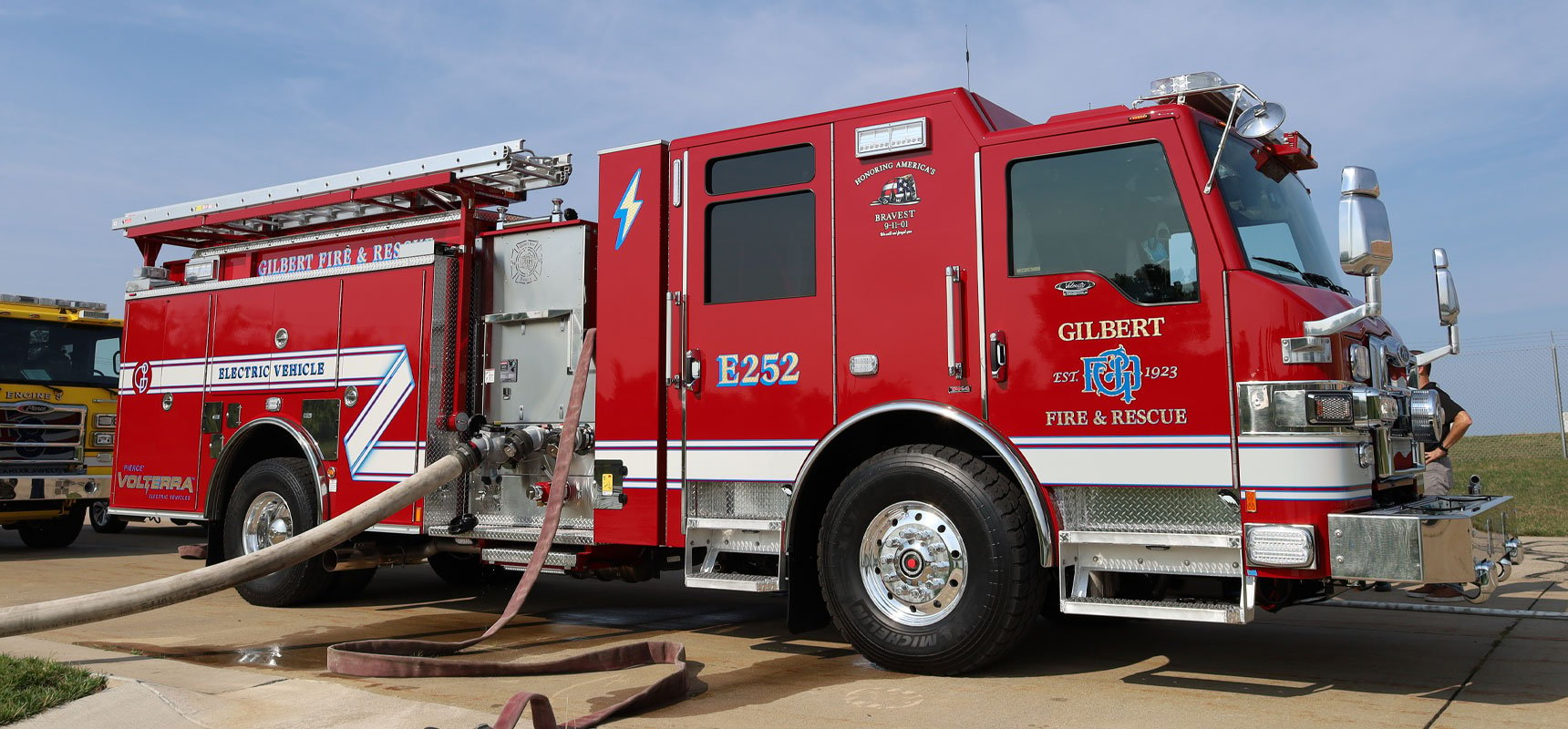 A red electric pumper truck with hoses attached and a blue sky in the background.