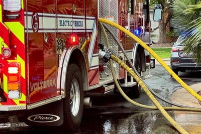 A red electric fire truck pumper is parked on the side of a road with hoses connected to the pump panel as it pumps water for emergency response. 