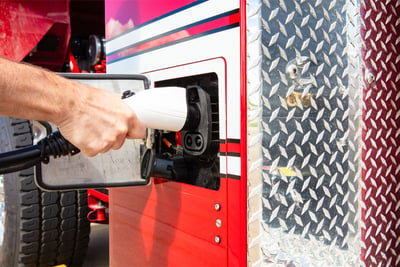 A hand is shown plugging a red electric fire truck into the power source for charging. 