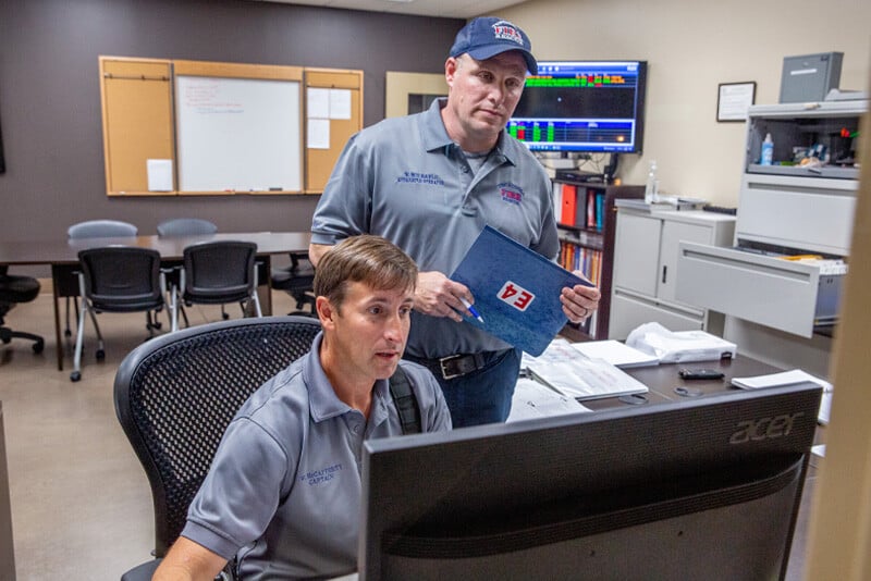 Two firefighters, one sitting and one standing, look at a computer monitor together in an office setting.  