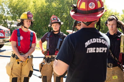 Madison Fire Department firefighters in helmets and hear gather listening to one firefighter lead training. 