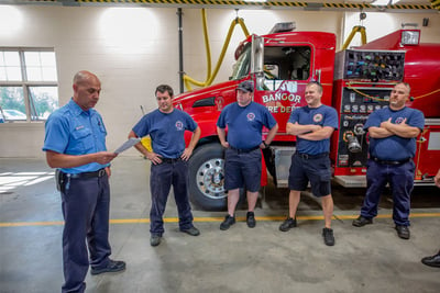 Members of the Bangor Fire Department stand alongside a red fire truck as the fire chief reads from a piece of paper in his hand. 