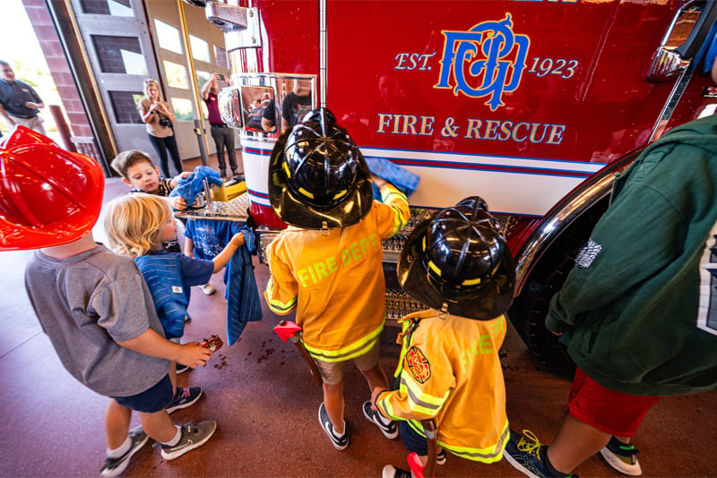 A group of children dressed in pretend firefighter gear are using blue cloths to polish a red fire truck at a community event inside a fire station.  