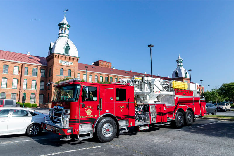 A red fire truck is parked in an asphalt parking lot with cars and a building in the background. 