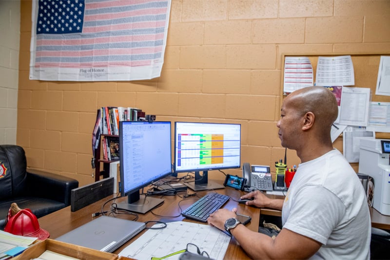 A man sits at a desk in a fire station working at a computer with two monitors. 