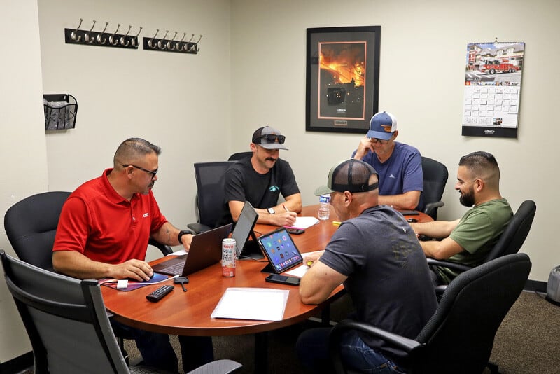 Five fire truck committee members sit around a table in a conference room looking at computer and tablet screens. 