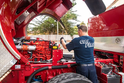 A fire truck mechanic with a blue shirt that reads ‘CAL FIRE’ works under a raised fire truck cab.