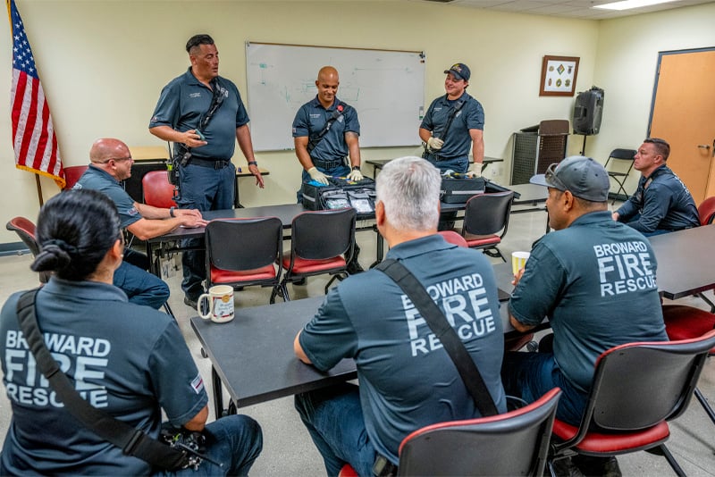 Firefighters in blue 'Edward Fire Rescue' uniforms sit in a classroom, facing three standing firefighters leading a discussion at the front.