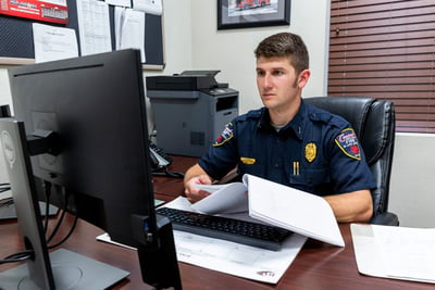 A firefighter in a blue uniform is seated at a computer desk with documents in his hands as he reads the computer screen. 