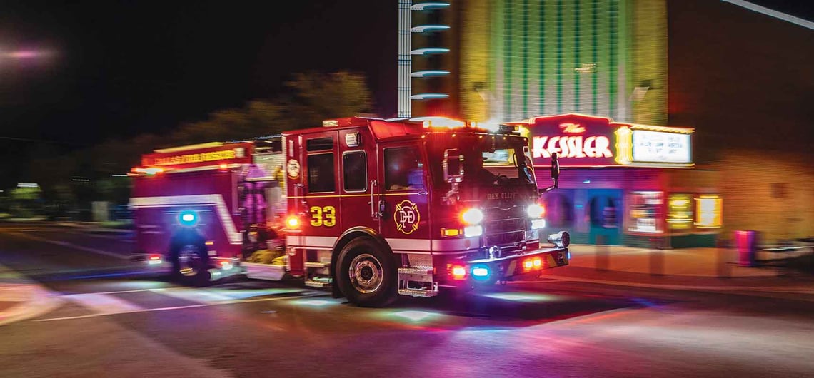 A fire truck is driving in the dark, illuminated with apparatus lights and the backdrop of a nighttime theatre scene.