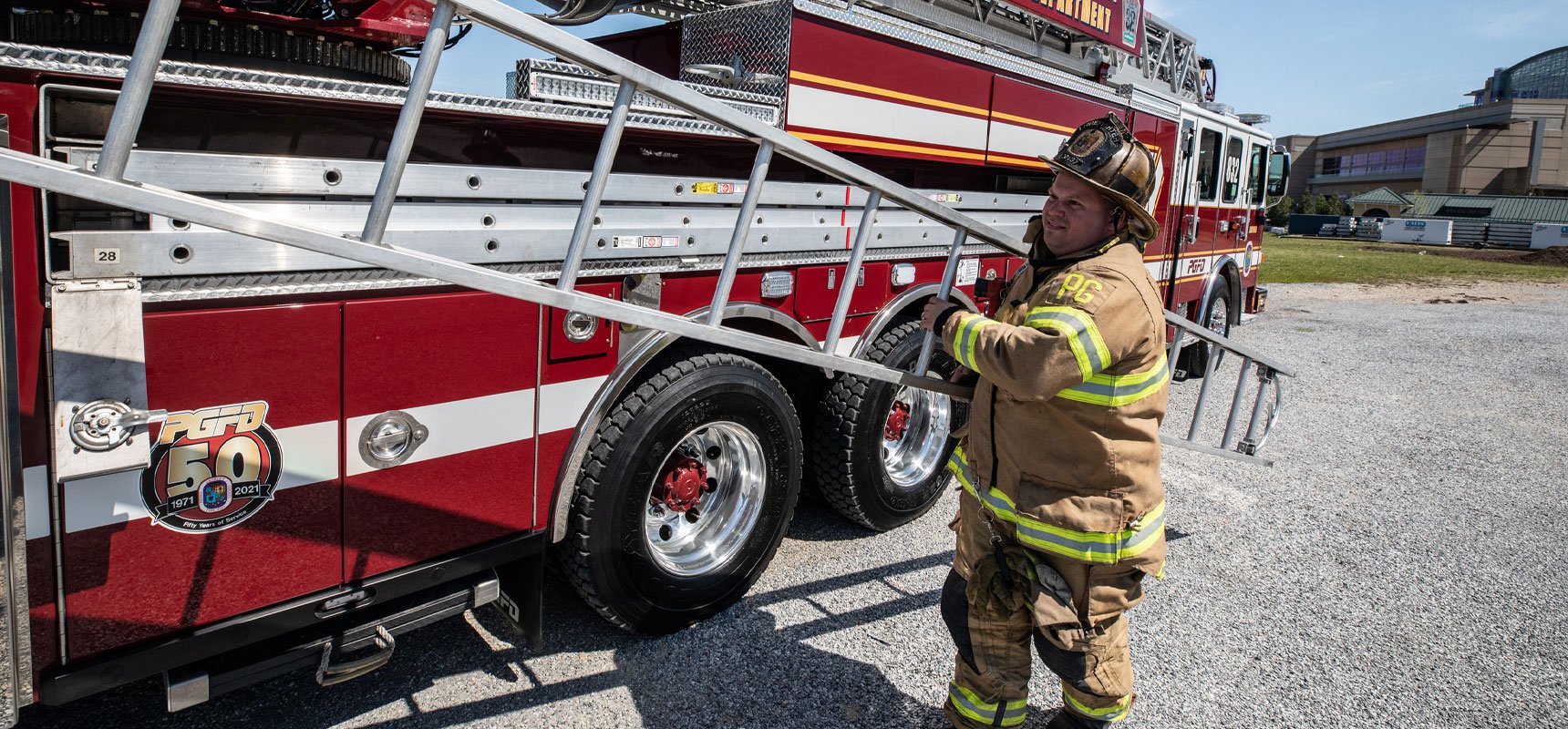 A firefighter in bunker gear pulls a ground ladder from an ergonomically positioned side compartment of a red Pierce apparatus parked on a gravel-covered area.