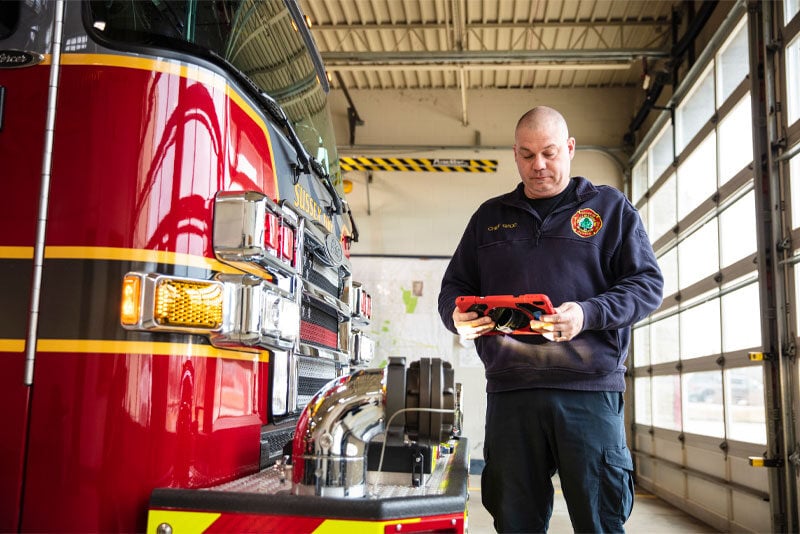 A firefighter looks at a table as he stands next to a red, black and yellow fire truck in a garage.