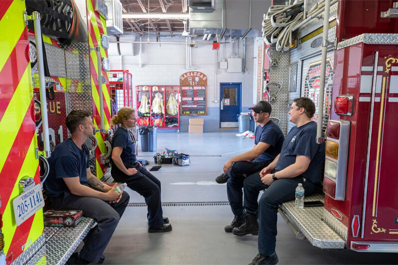 Four firefighters in casual blue uniforms sit facing one another on the bumpers on two adjacent firetrucks inside a fire station. 