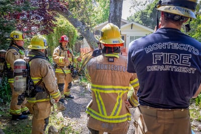 Several Montecito Fire Department firefighters with helmets and gear stand in a semi-circle outside of a home as one firefighter leads a discussion. 