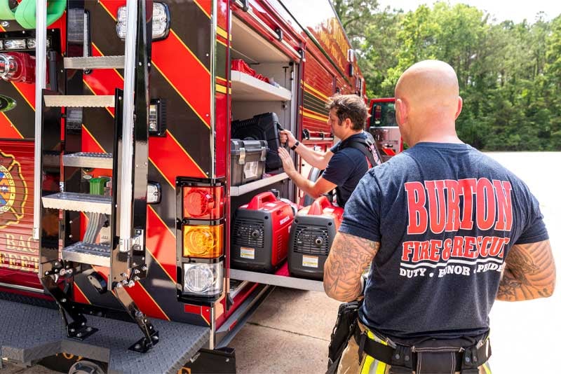 Two Burton Fire and Rescue firefighters are removing gear from a compartment on a red and black fire truck. 