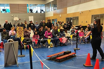 A group of people site on bleachers in a gym where a female is presenting various firefighting techniques.