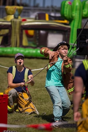 A kid wears a firefighters jacket over his teal shirt while pulling on a rope with a female firefighter in the background.