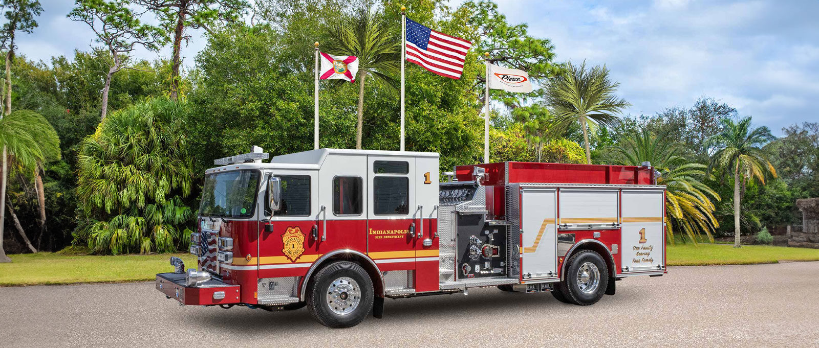 The red and white Indianapolis Fire Department pumper is parked on an asphalt surface with three flag poles and green vegetation in the background. 