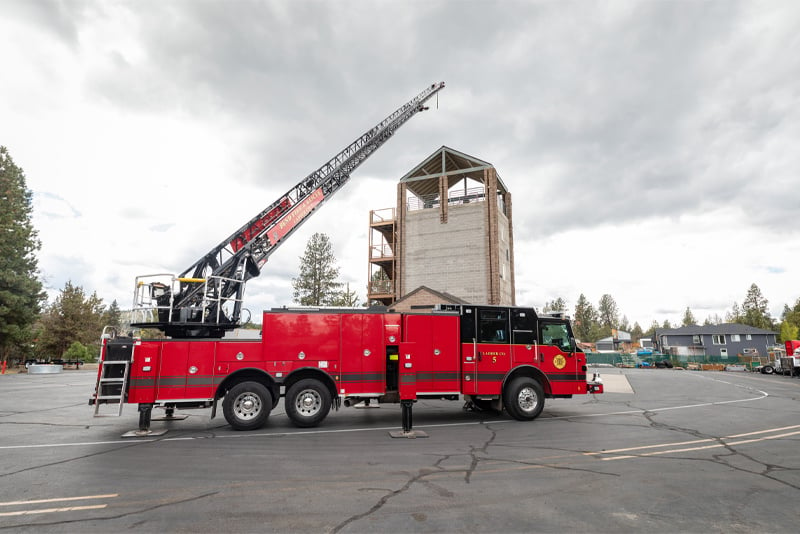 A red aerial apparatus has its stabilizers set and the ladder extended in a fire department parking lot during training exercises. 