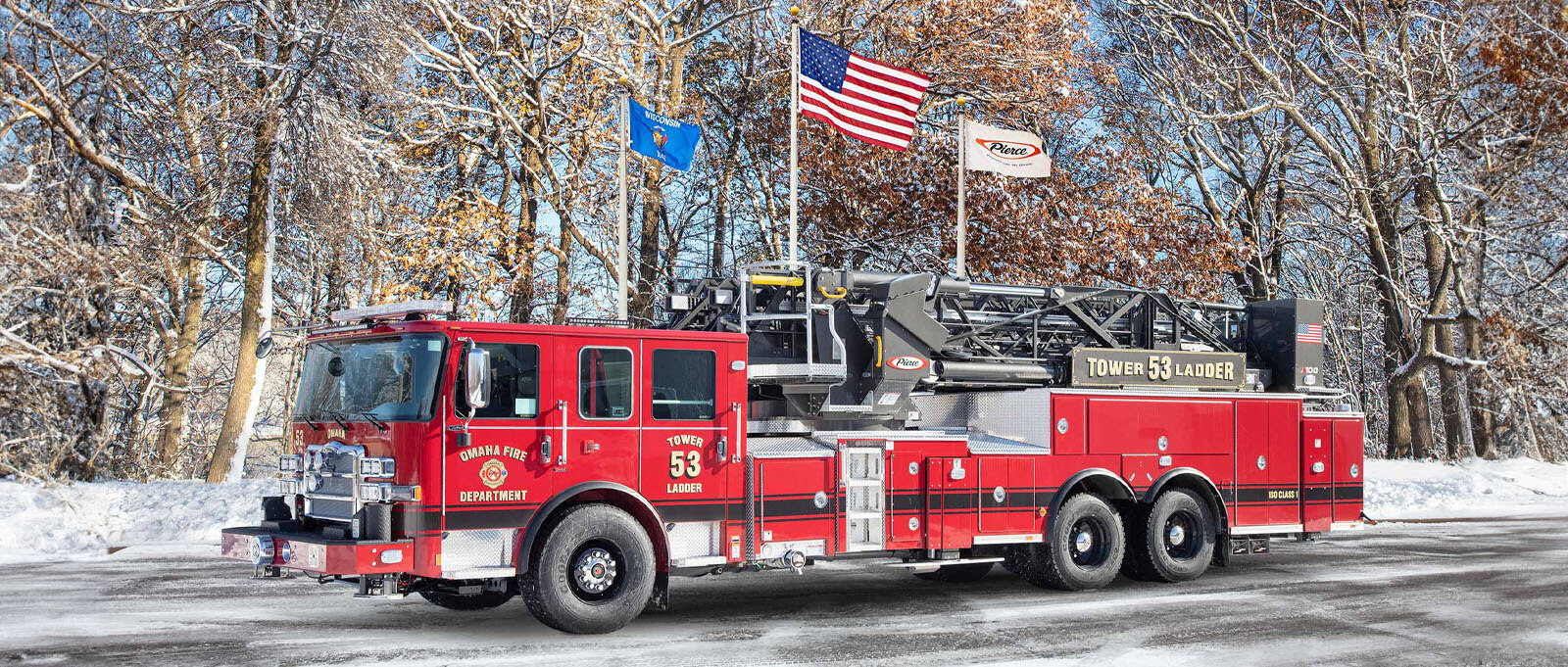 The red Omaha Fire Department aerial fire truck is parked on an asphalt surface with three flag poles, snow and trees in the background. 