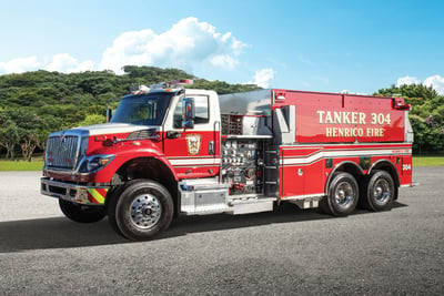 A red Pierce commercial dry side tanker is parked on an angle with green foliage and three flags in the background.  