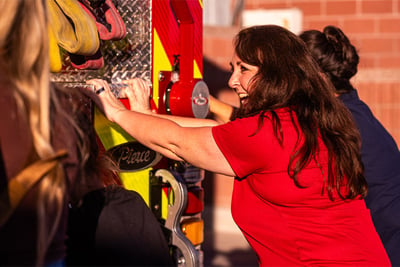 A smiling woman with brown long hair in a red shirt has two hands on an apparatus as she helps push it in a traditional push in ceremony. 