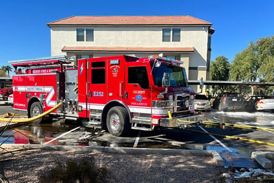 A red electric fire truck is parked in front of a residential home with hoses deployed pumping water. 