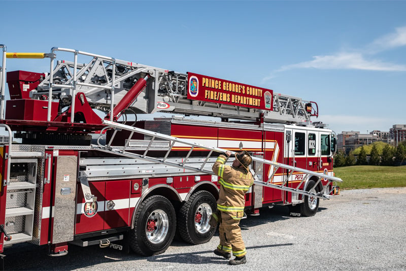 A firefighter in bunker gear is removing a ladder from the side of a red aerial apparatus which is parked on asphalt in an industrial area. 