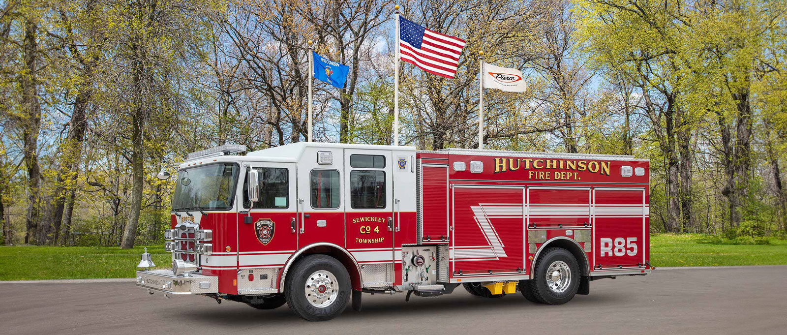 A red Sewickley Township Volunteer Fire Company & Relief Association pumper is on an asphalt surface with three flag poles and trees in the background