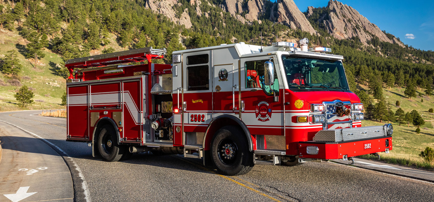 A red and white Pierce fire truck sitting on a winding pavement road with mountains and trees in the background.