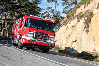 A red fire truck, with a '21' on the grill, driving down a hilly winding road, with a mountain on the side.