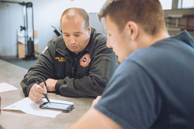 Two fire truck committee members are seated at a table, with one using a pen to point to a phone screen and papers resting on the tabletop. 