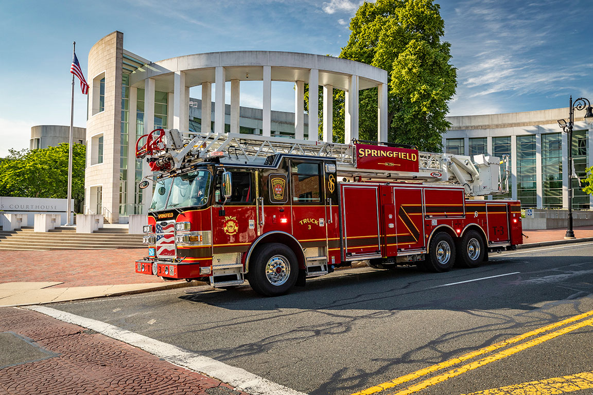 An Arrow XT 100' Heavy-Duty Low-Profile Steel Aerial Ladder driving in front of a courthouse with white pillars and an American flag.